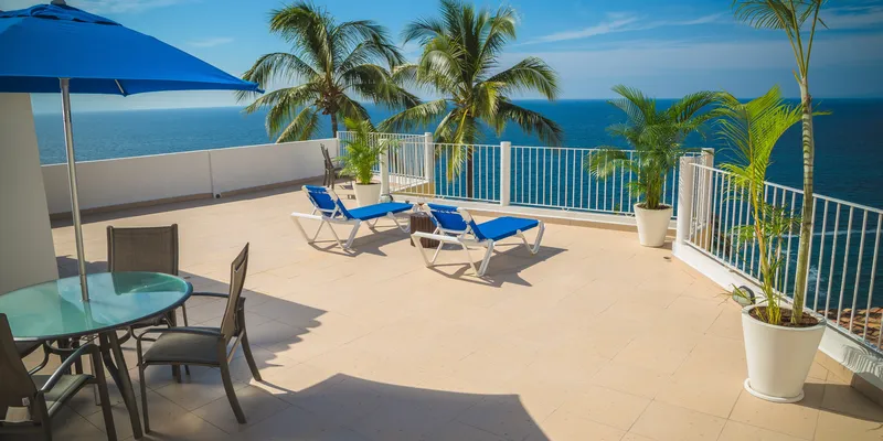 Terrace with table, chairs, blue umbrella, plants, sunbeds, and ocean view with palm trees 
