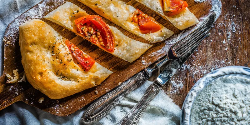 Bread with tomato on wooden platter with fork and knife and flour