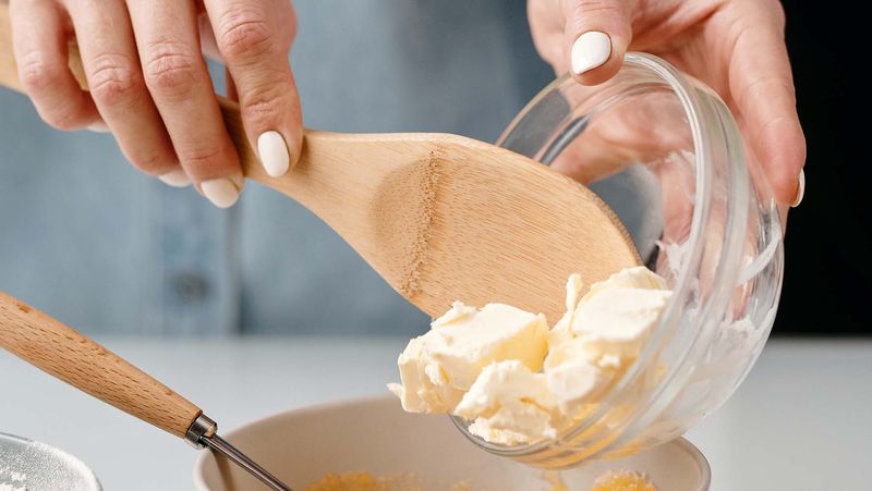 Hands holding a wooden spoon emptying butter into a mixing bowl
