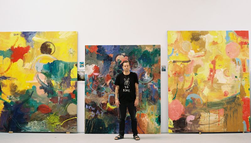 Artist stood before three large scale canvases behind him with colourful patterns on