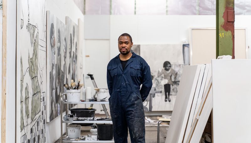 artist wears dark blue overalls and stands with hands behind his back in his studio, with empty canvases to his left and a selection of his artwork on the wall to his right