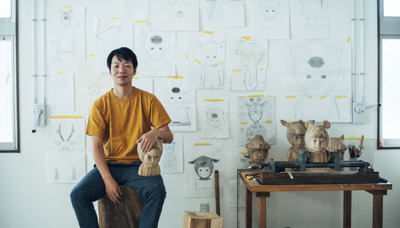 Satoru Koizumi sat on a block of wood with his arm resting on a small wooden sculpture placed on his knee, in front of a wall filled with pencil sketches on paper