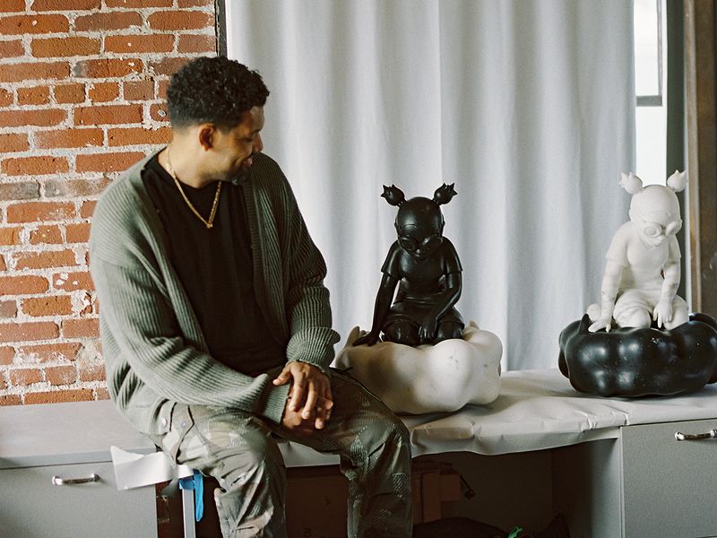 Artist in khaki green cardigan sitting on a table next to two of his marble sculptures