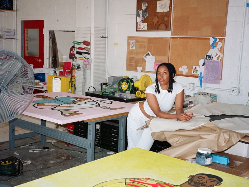 Tschabalala Self leaning on a table in her studio, a large yellow painting is visible in the foreground