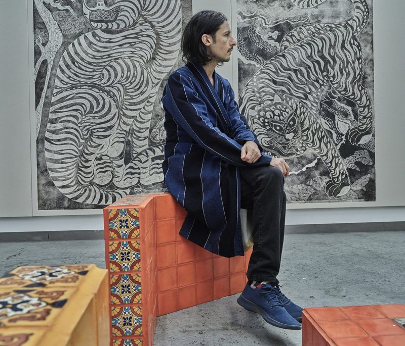 Kour Pour sat on a rust-coloured block in his studio with two monochrome works behind him