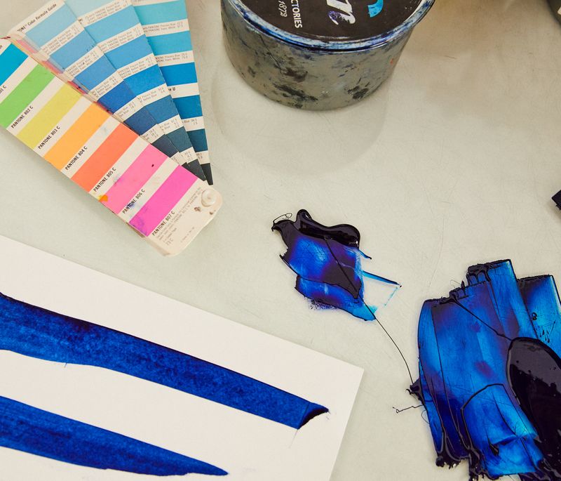 a table with strips of various colours on, next to a piece of paper where various marks have been made with blue paint