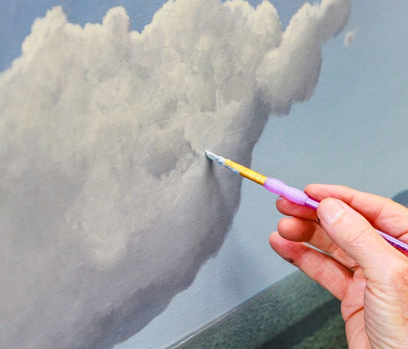 purple paintbrush dipped in white paint, adding details to a white cloud that has been painted on the canvas