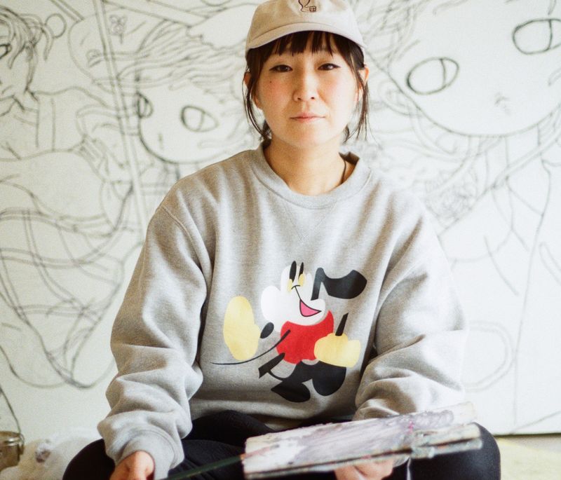 Aya Takano sitting cross-legged on the floor with a paintbrush in her hand