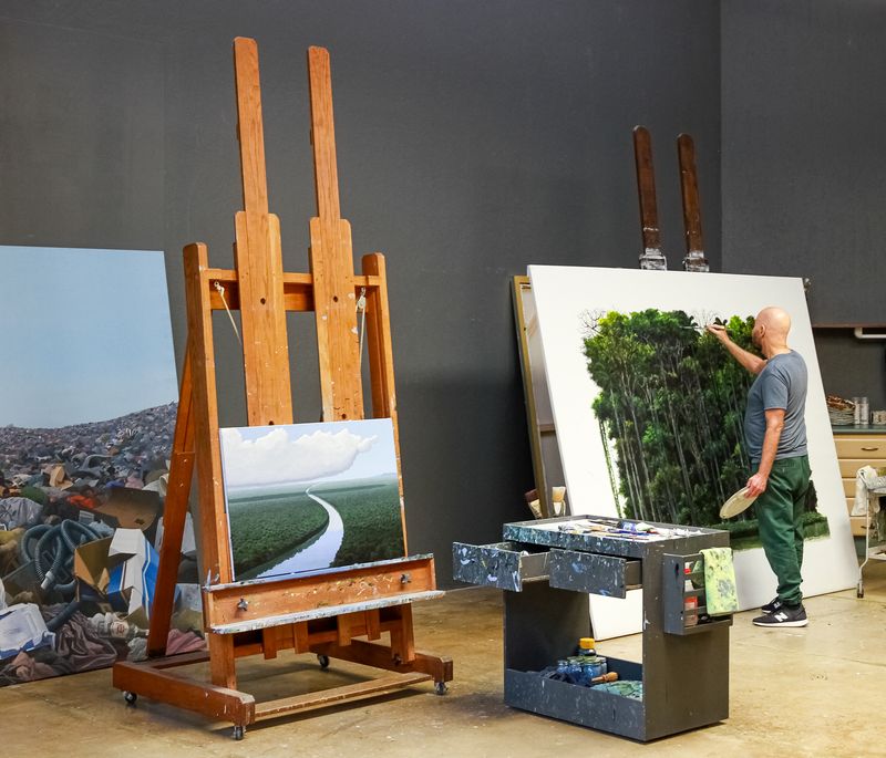 artist stood in front of a large white canvas with a green forest painted on it with trays of paints to each side of him, as well as an easel and a large painting of landfill 