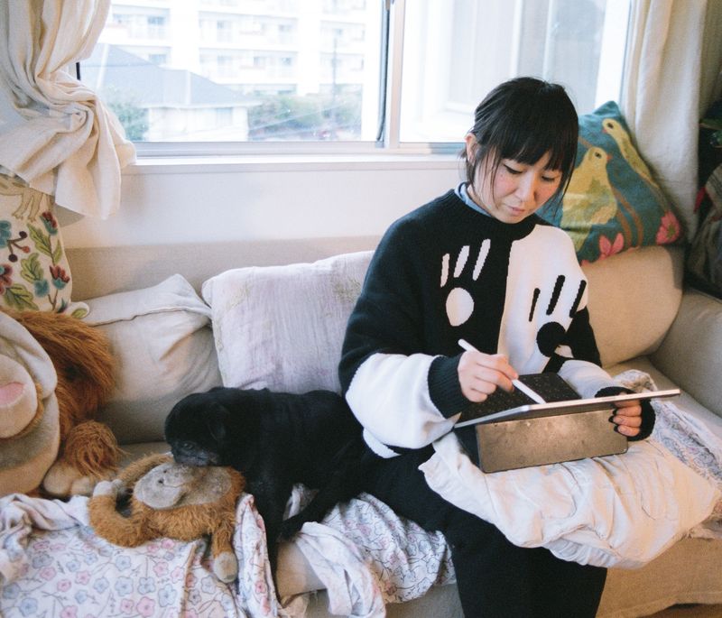 Aya Takano sitting on a sofa with her pug and drawing on her tablet