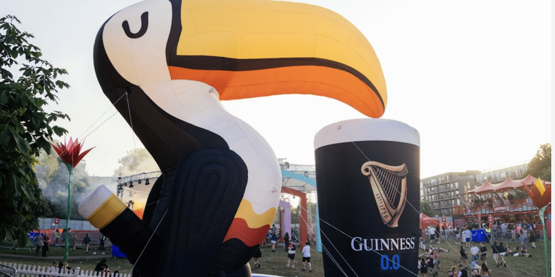 Inflatable toucan and a large Guinness beer can in a festival setting with people and tents in the background.
