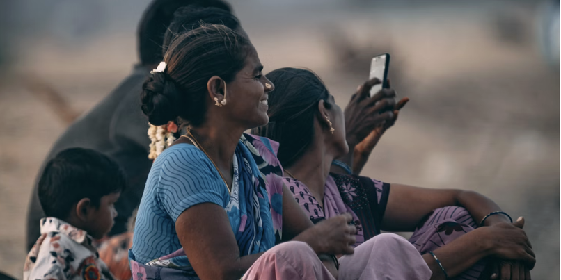 A group of women and a child sit outdoors, all are relaxed. One woman is holding a smartphone. The scene is informal and candid.