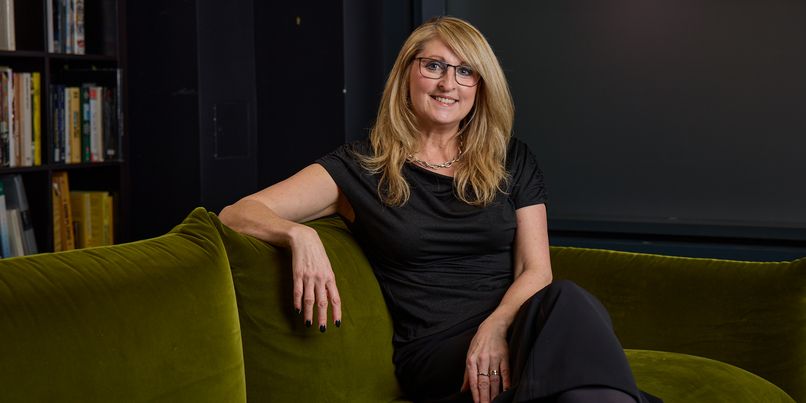 Anne Stagg, CEO of Digitas UK with long blonde hair and glasses sits relaxed on a green sofa, wearing a black outfit, with bookshelves in the background.