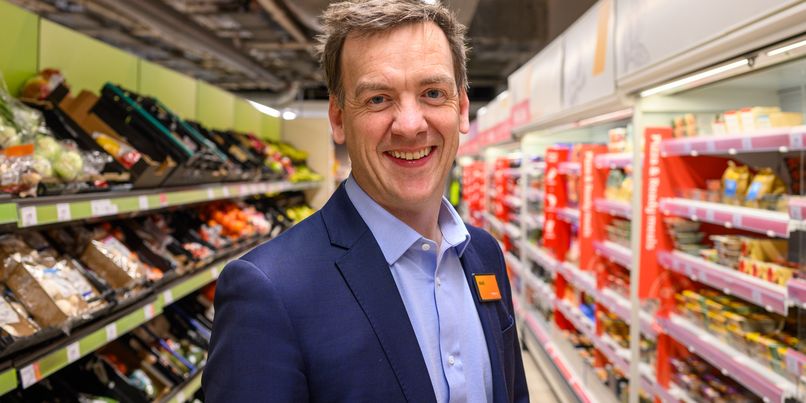 Mark Given, Sainsbury's marketing chief in a suit stands in a grocery store aisle stocked with fresh produce and packaged goods.
