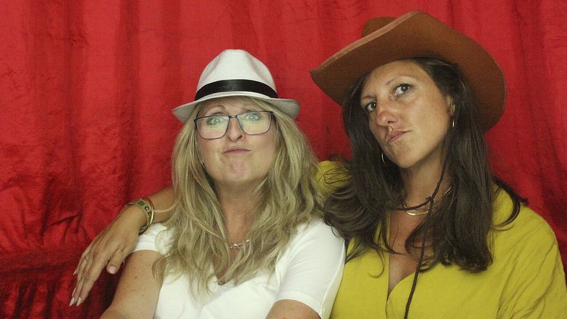 Two women pose in front of a red curtain, one wearing a white fedora and glasses, the other a brown cowboy hat, both smirking playfully.
