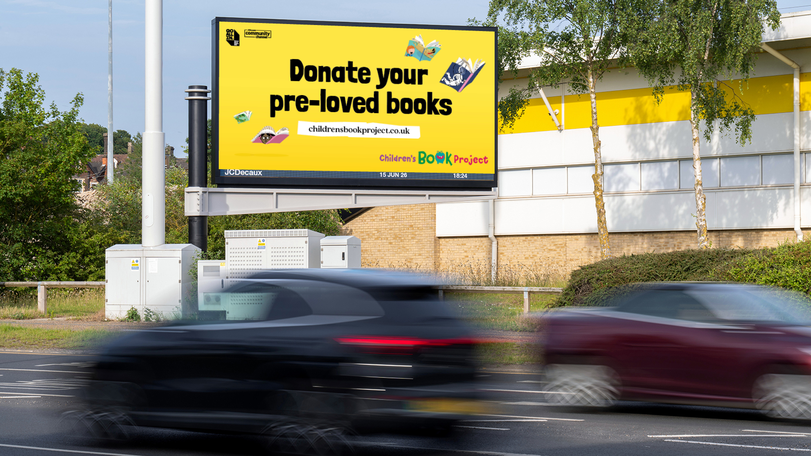 A roadside billboard reads "Donate your pre-loved books" for the Children's Book Project, with cars passing by.