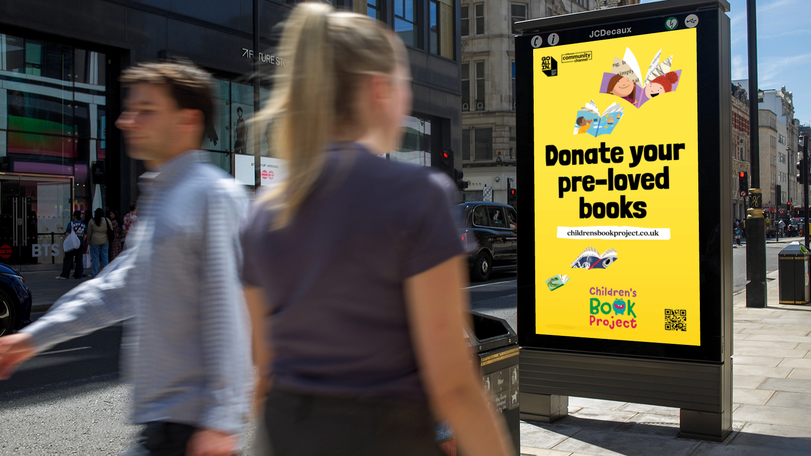 Street view with pedestrians walking past a bright yellow advertisement encouraging the donation of pre-loved books for a children's book project.