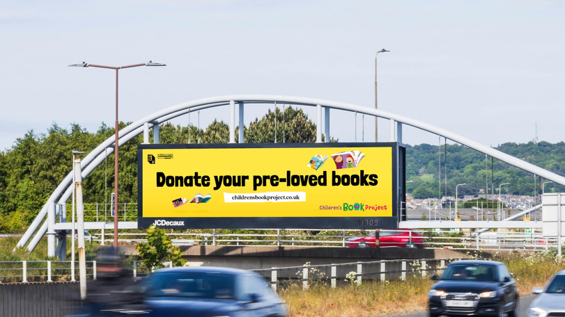 A highway billboard reads, "Donate your pre-loved books" with the website childrensbookproject.co.uk. Cars pass by below.