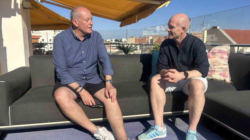 Two people sitting on a rooftop patio couch, engaged in conversation under a yellow awning, with a sunny sky in the background.