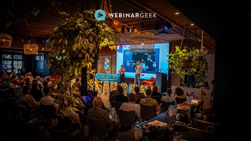 A lively conference room with a speaker on stage, audience seated, and hanging plants in the foreground.