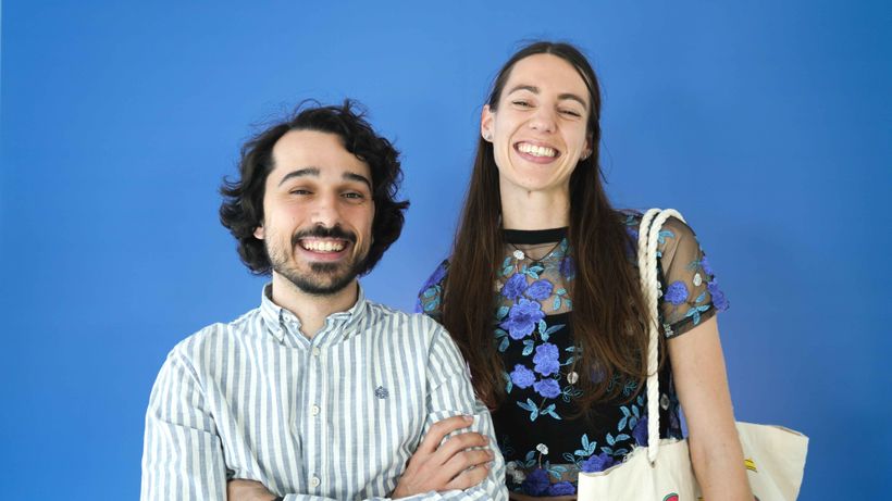 Two people smiling brightly against a blue background; one wears a striped shirt, the other a floral top.