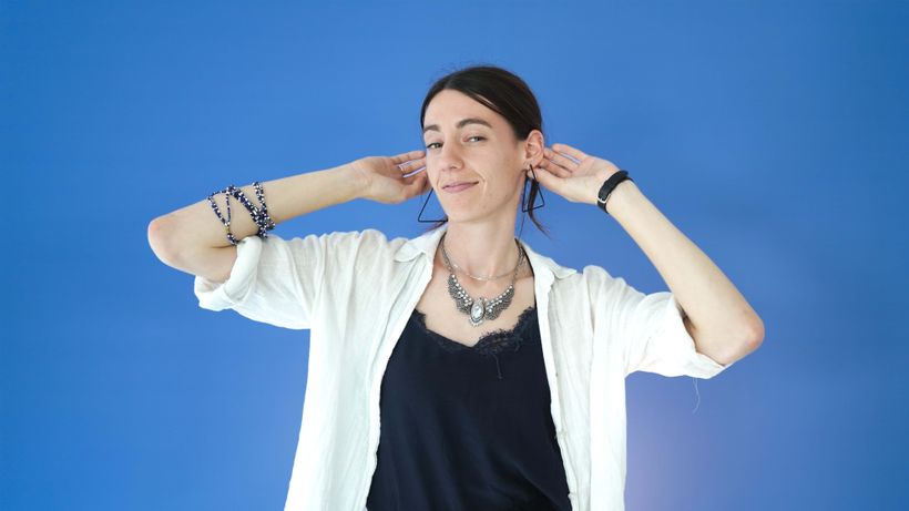 A woman poses against a blue background, wearing a white shirt, black top, layered necklaces, and holding her hair with both hands.