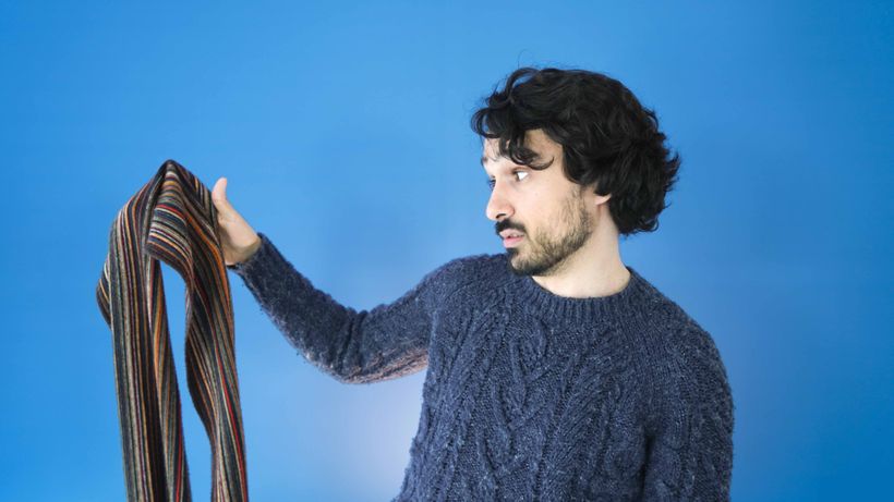 A man in a blue sweater holds a colorful striped scarf against a bright blue background.