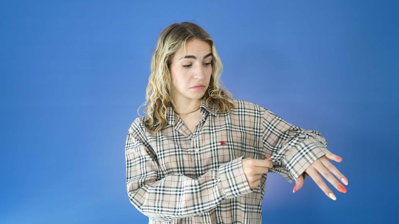 A person with wavy blond hair and colorful nails points to their wrist, wearing a plaid shirt against a blue background.