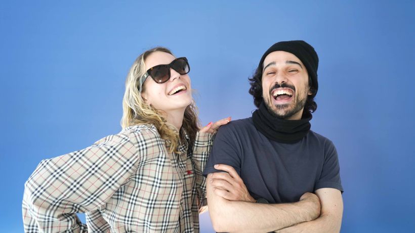 A man and woman laugh together against a blue background. The woman wears sunglasses and a plaid shirt, while the man wears a beanie and scarf.