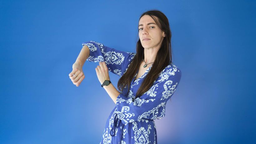 A woman in a blue patterned dress poses with her arm bent, against a solid blue background. She wears a black watch and necklace.