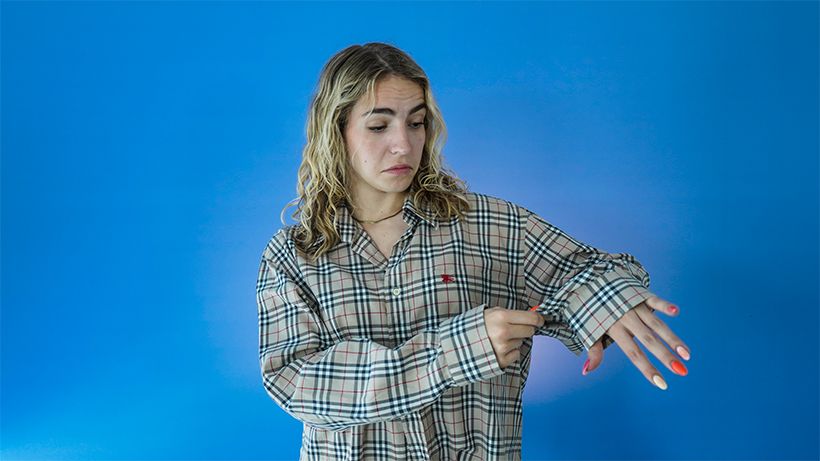 Person in a plaid shirt looking at their wrist with a puzzled expression, against a blue background. Brightly painted nails.