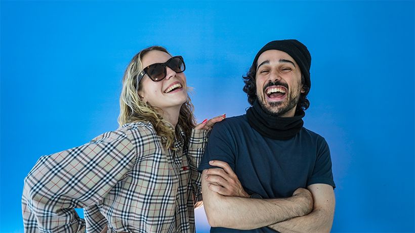 A woman in sunglasses and a man in a beanie laugh joyfully against a bright blue background.