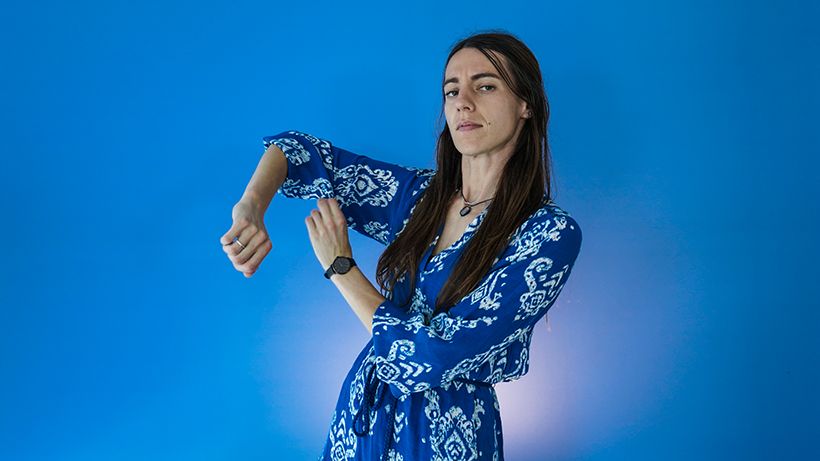 Person in a blue patterned dress rolling up their sleeve against a bright blue background.