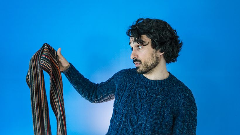 Man in a blue sweater holds a colorful striped scarf against a blue background, looking at it thoughtfully.