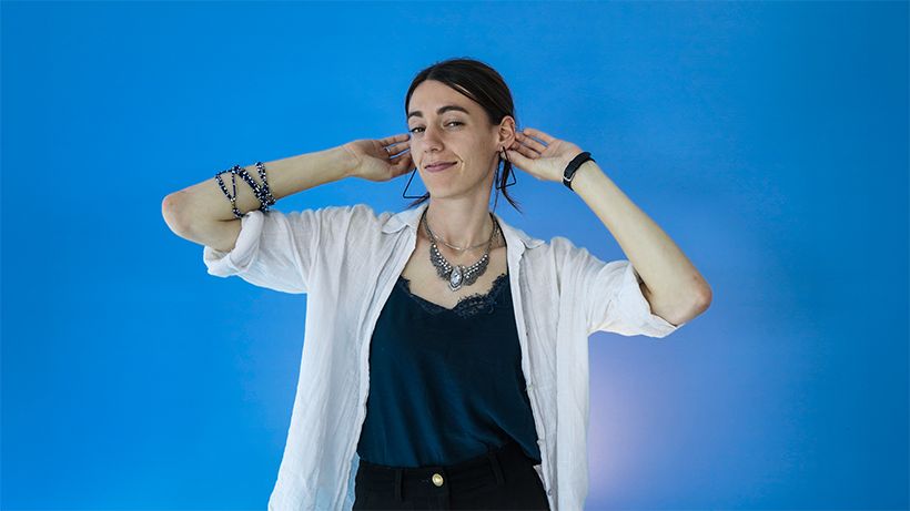 Woman in a white shirt and jewelry playfully poses against a blue background, with hands near her face, smiling slightly.