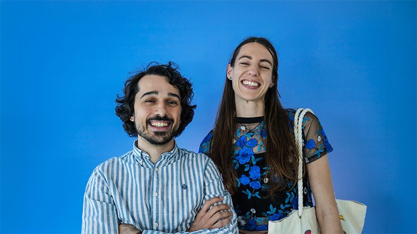 A smiling man and woman stand together against a solid blue background. The man wears a striped shirt, and the woman wears a floral top and holds a tote bag.