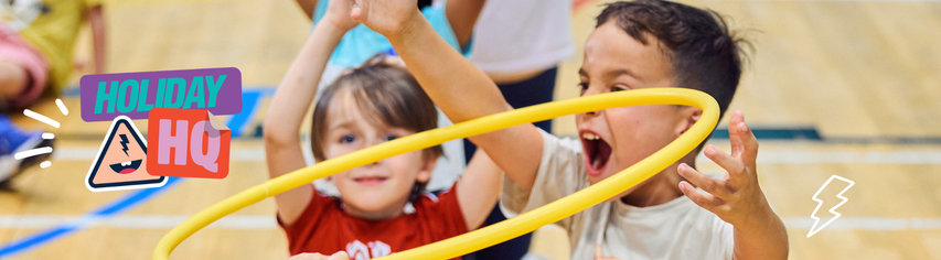 Two young children excitedly play with a yellow hula hoop indoors. Graphic text says "Holiday HQ" with colorful icons.