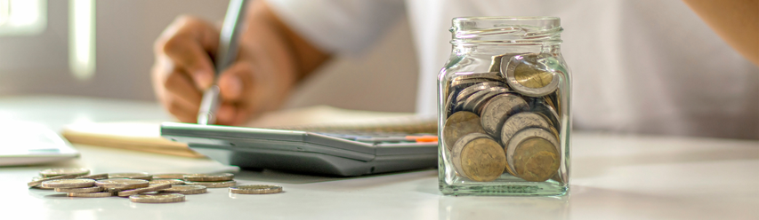 Person calculating with a notebook and calculator, next to a glass jar filled with coins on a table.