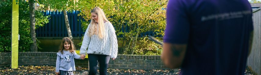 A woman and a young girl hold hands, walking outside near a brick wall and trees, with a person in a purple shirt in the foreground.