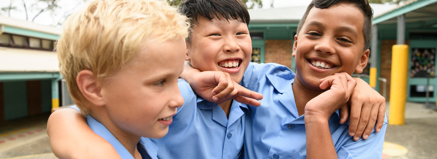 Three smiling boys in blue uniforms, embracing each other outdoors at school.