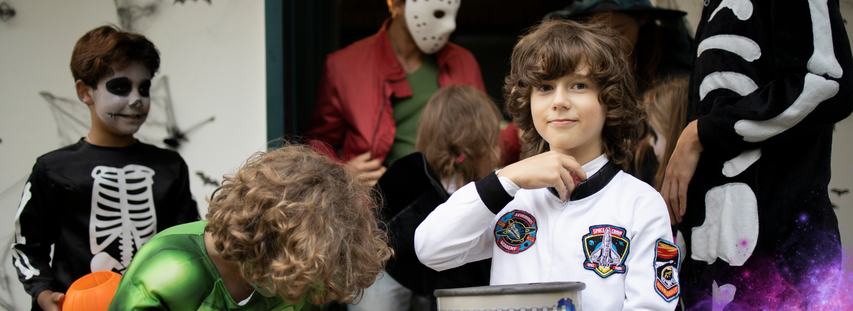 Children in Halloween costumes, including a skeleton, astronaut, and superhero, gather with candy buckets and decorations in the background.