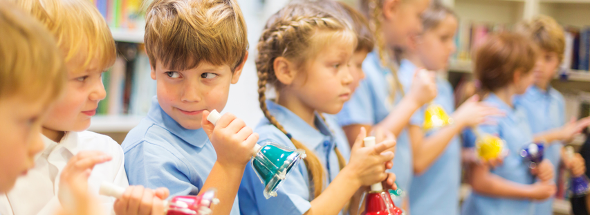 Children in light blue shirts playing colorful handbells, standing in a line. Focus is on two children holding green and red bells.