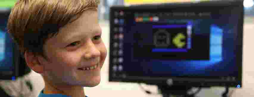 Smiling boy in a blue shirt sits in front of a computer displaying a pixelated game character on the screen.