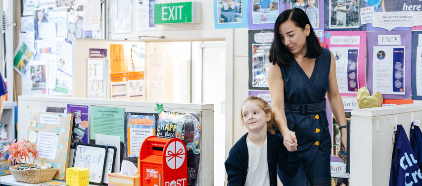 A woman holds a young girl's hand in a colorful classroom, decorated with posters and a visible exit sign above.