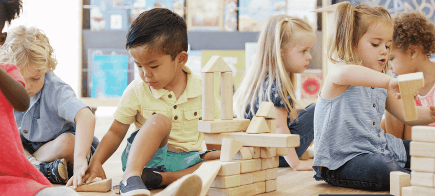 Children sitting on the floor, playing and building with wooden blocks in a classroom setting.