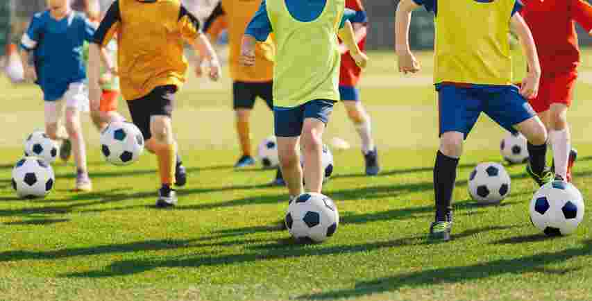Children in brightly colored sportswear practicing soccer, each dribbling a ball on a green field.