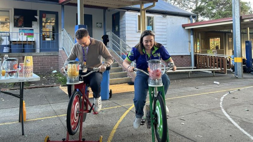 Two people pedal stationary bikes with blenders attached, making smoothies outdoors at an event.