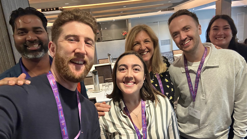 A group of six people smiling and posing for a selfie indoors, wearing conference lanyards.