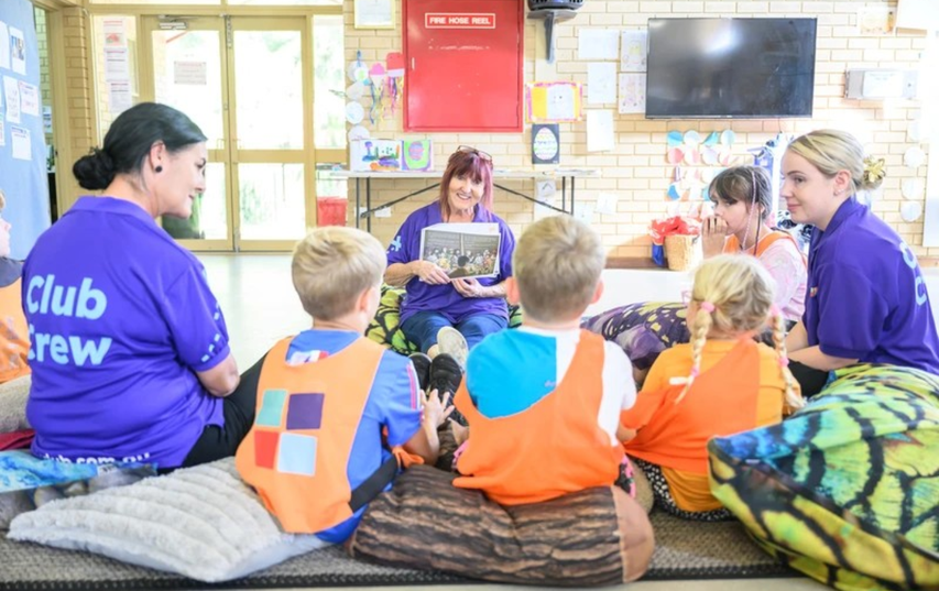 A group of children and staff in purple shirts, labeled "Club Crew," sitting on cushions during a storytelling session in a classroom.