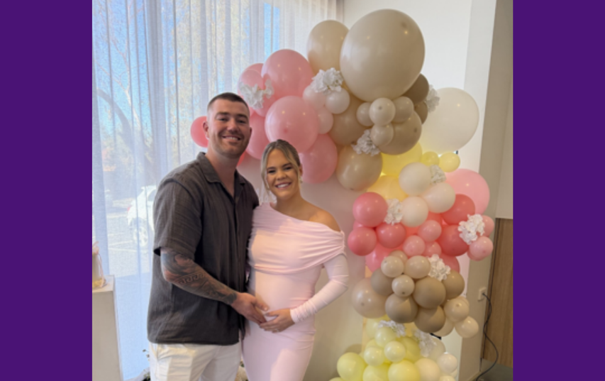 A smiling couple stands in front of a colorful balloon display, with the woman in a light pink dress holding her pregnant belly.